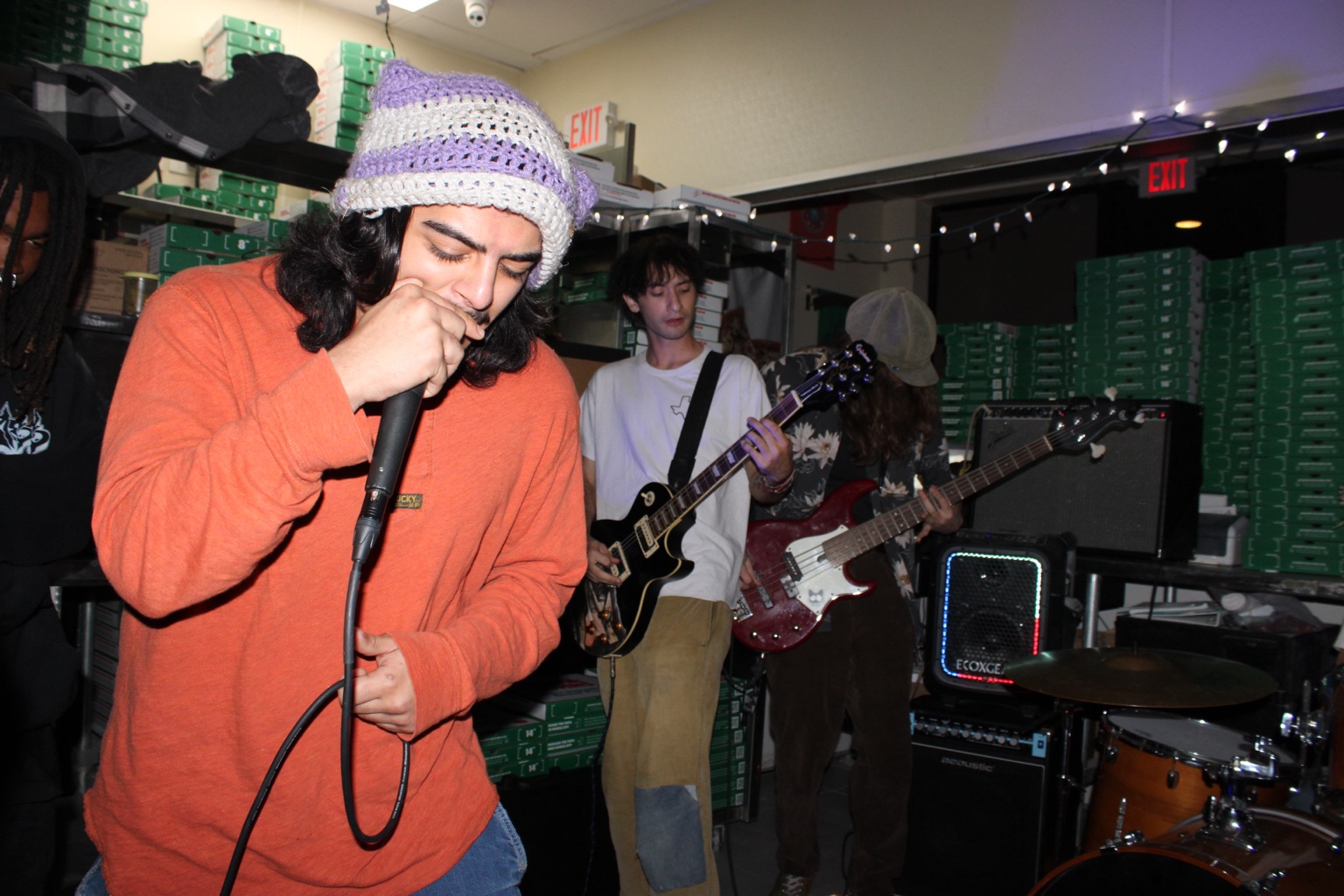 A vocalist in an orange sweatshirt performs with guitarists in an intimate room lined with green boxes.